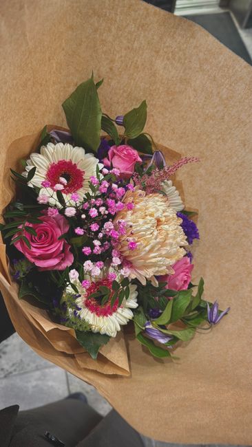 A bouquet of fresh flowers arranged in a natural style, featuring pink roses, white daisies with pink centers, creamy peonies, and small purple and pink filler flowers, accented with green leaves. The