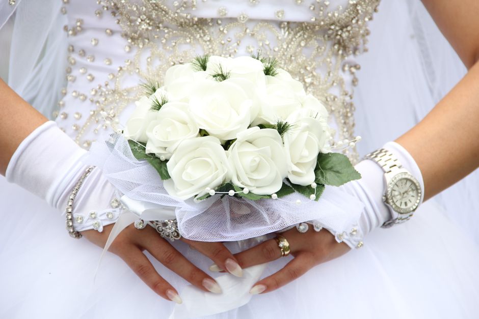 A bride in a white lace wedding dress is holding a floral bouquet, featuring pale pink and cream roses, white orchids with pink accents, and small white filler flowers, accented with lush green fern l